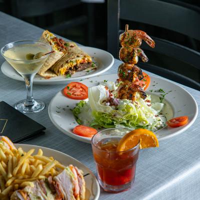 A table set with a wrap, a club sandwich with fries, wedge salad with shrimp, and two cocktails.
