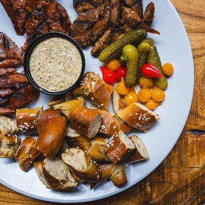 Appetizer platter with sliced sausage, soft pretzel bites, whole grain mustard, pickled veggies.