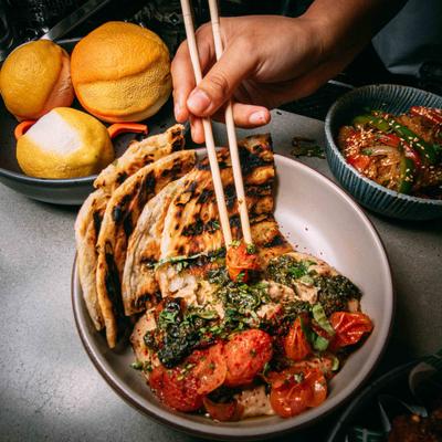 Person using chopsticks over grilled flatbread and tomatoes.