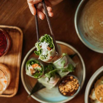 A hand with chopsticks lifting a spring roll from a plate.