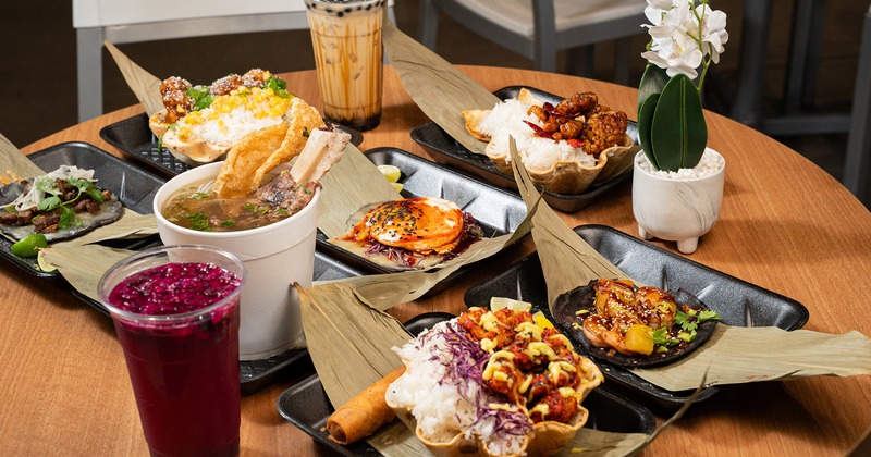 Various Asian-inspired dishes, including tacos, bowls, soup, and drinks, are arranged on a table.