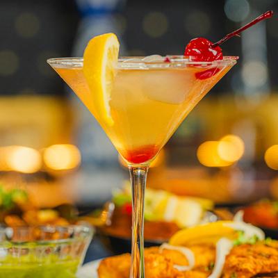 A classic cocktail with a lemon twist and cherry garnish sits on a table with food.