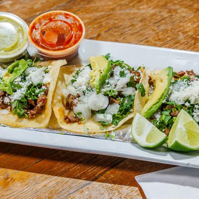 Street tacos topped with cotija cheese and avocado, served with two cups of salsa.