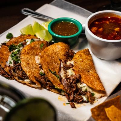 Queso birria tacos served with birria broth, salsa, and lime.
