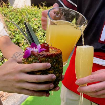 Two hands holding cocktails, one in a pineapple with a flower and the other in a flute.