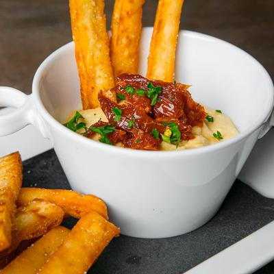 A bowl of creamy dip topped with sun-dried tomatoes and naan bread sticks.