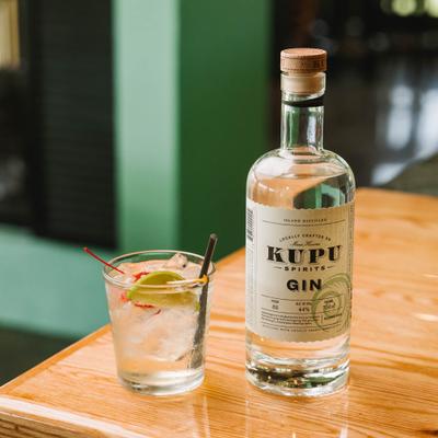 Cocktail garnished with lime and cherry sits beside a bottle of gin on a table.