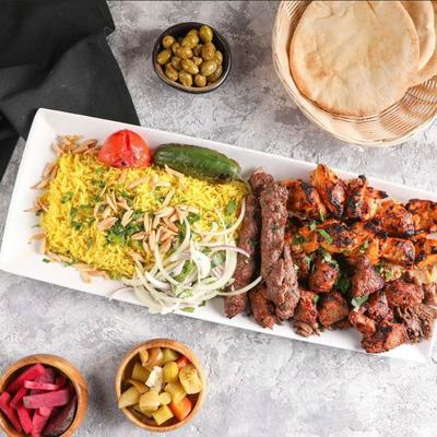 A platter with grilled meats, yellow rice, and onions, sides include pickles, olives, and pita.
