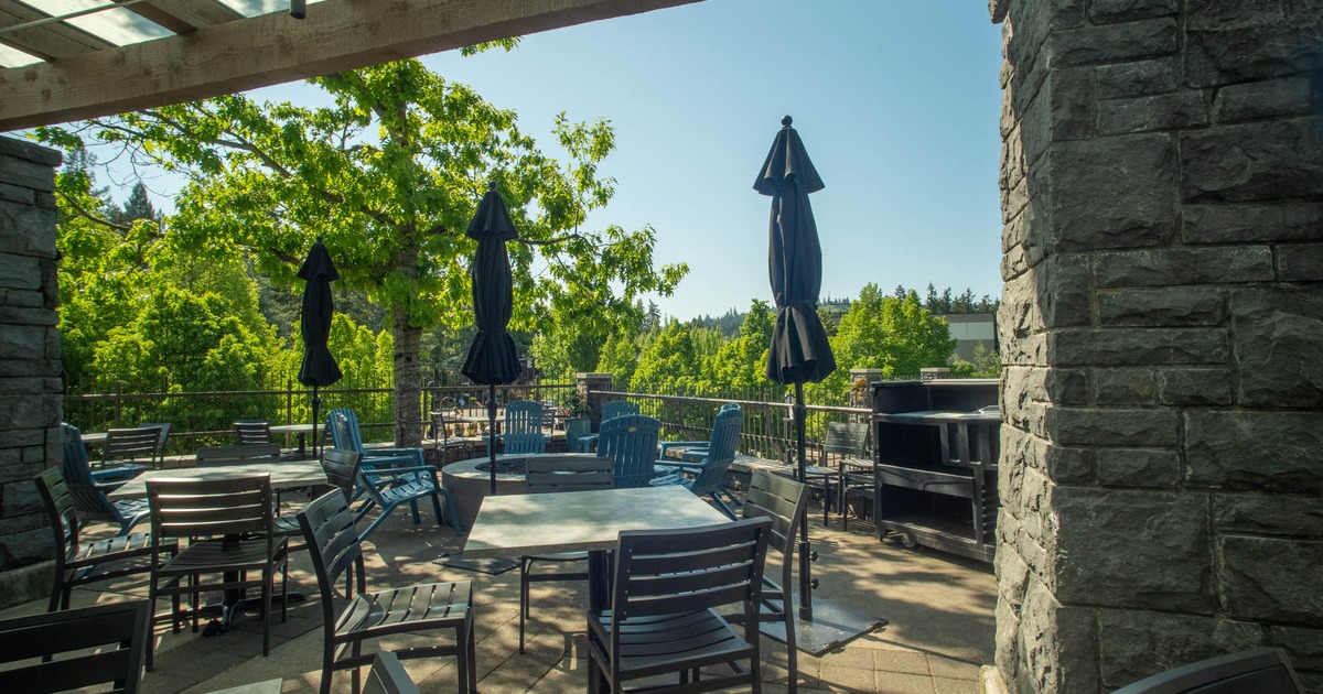 Sunlit outdoor patio with stone walls, tables, and black umbrellas, surrounded by trees