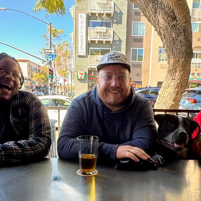 3 smiling people sitting at a bar with a street behding them with drinks and Bruno the black and white adorable dog.