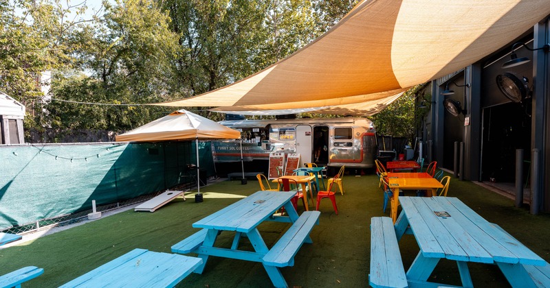 Outdoor dining area with wooden blue tables and benches and shade sails above.
