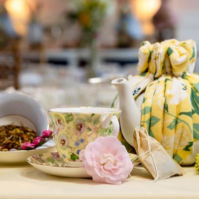 A floral teacup surrounded by a teapot in a tea cozy, loose tea, and a flower.