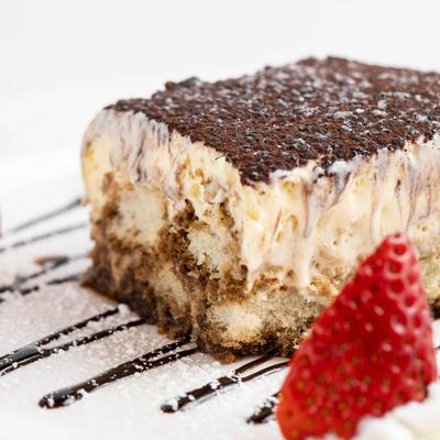 Tiramisu topped with cocoa powder, on a plate with strawberry garnish and chocolate sauce drizzle.