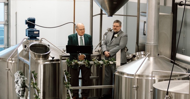 Two men in suits look over brewing equipment on an elevated metal platform.