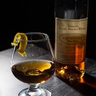 A glass of bourbon with a lemon twist on a wooden table, paired with an open bottle beside it.