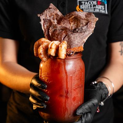 Gloved hands holding a Michelada drink garnished with grilled meat and shrimp.