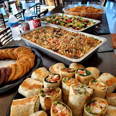 Catered spread featuring sliced wraps, foil trays of pasta and a vegetable salad.
