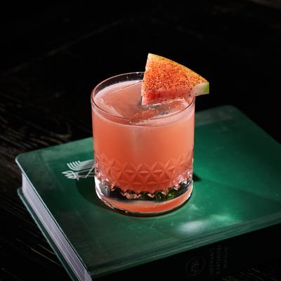 The Devil Knows, garnished with a chili-dusted watermelon slice sits on a closed green book.