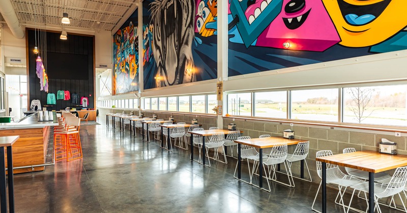 Interior, lined up tables by a window side opposite the bar, large mural art