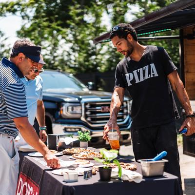 Three people at an outdoor pizza stand with assorted pizzas.