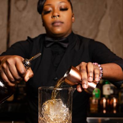 Bartender pouring a drink from a copper jigger into a mixing glass filled with ice.