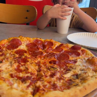 A happy, curly-haired child sits at a table behind a large pepperoni pizza.