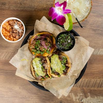 Street tacos with salsa, rice, beans, and margarita on wooden table.