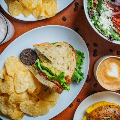 Sandwich with a side of chips and a cookie, served alongside other dishes and a coffee.