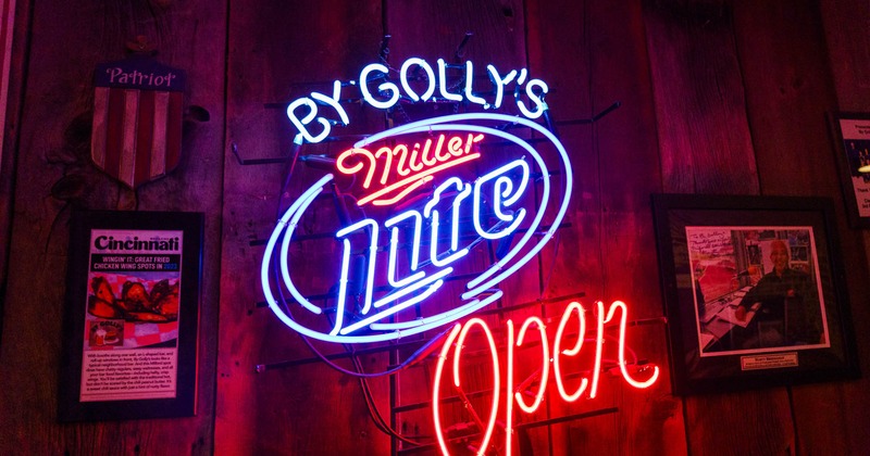 A neon sign reading By Golly's Miller Lite Open in blue and red colors on a wooden wall