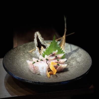 Sliced raw fish sashimi arranged on a dark ceramic plate with shiso leaf garnish.
