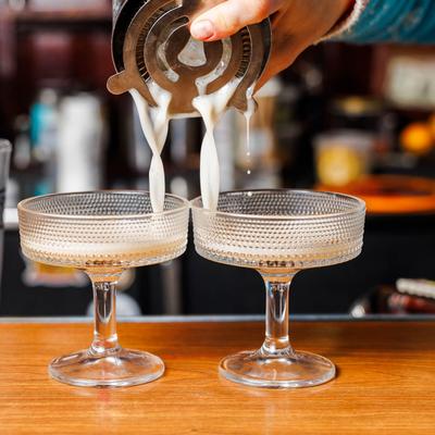 A bartender pouring a creamy cocktail from a strainer into two textured coupe glasses.