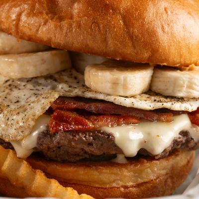 A close-up of a burger with bacon, cheese, banana slices, and a fried egg.