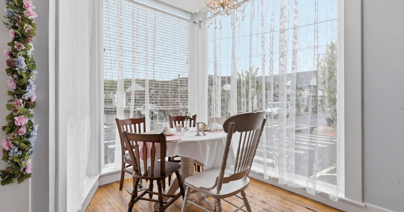 A bright cafe corner with a round table, wooden chairs, chandelier, lace curtains, and floral decor