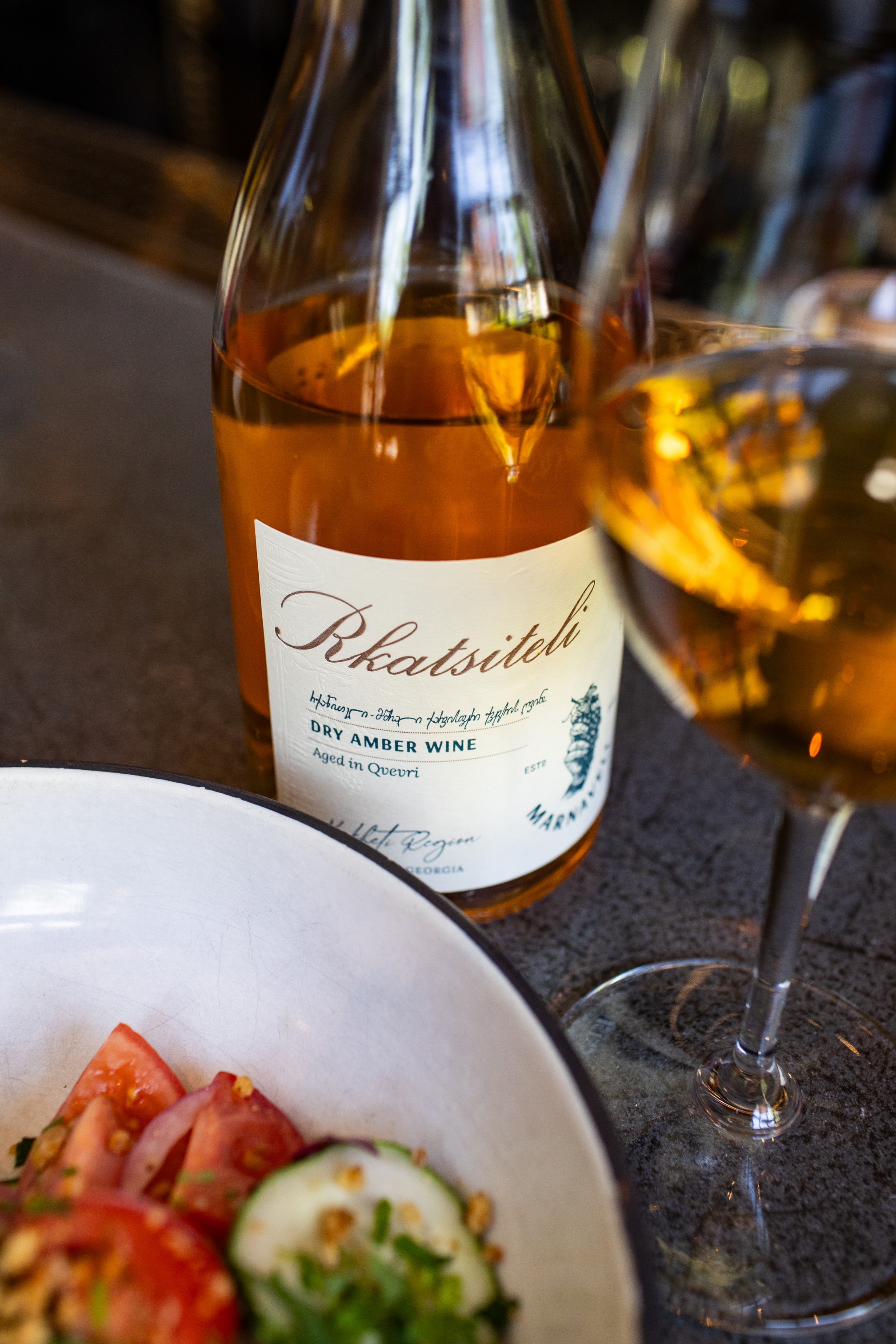 a bottle of golden 'amber' rkatsiteli wine from the Republic of Georgia beside a glass of the wine, next to a plate