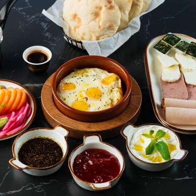 Baked eggs in a clay dish with flatbread, cheese, dips, sliced meats, and pickled vegetables.