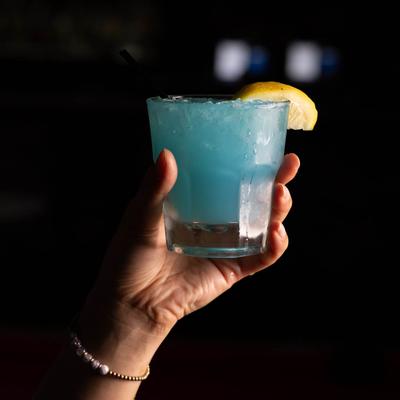 A hand holding an iced blue cocktail with a lemon wedge.