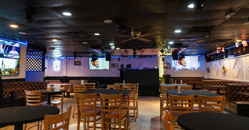Interior of a bar with black tables, wooden chairs, booths, TVs, and a central stage