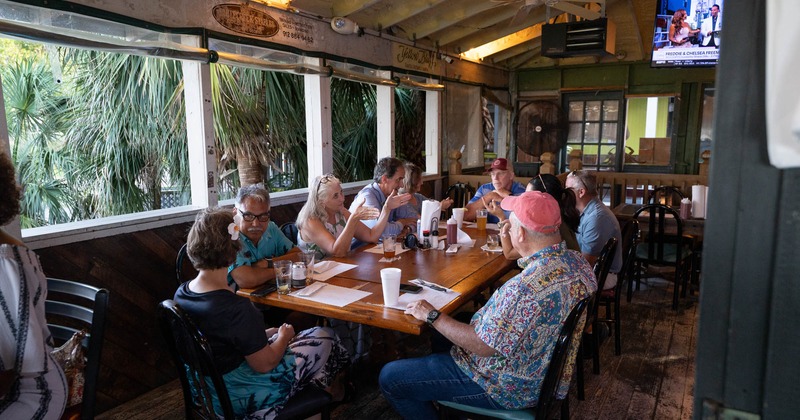 A group of people enjoy a meal at a wooden table in a casual, open-air restaurant