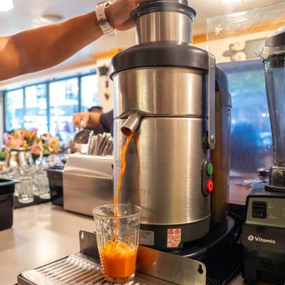 A hand pouring fresh orange juice from a stainless steel juicer into a glass.