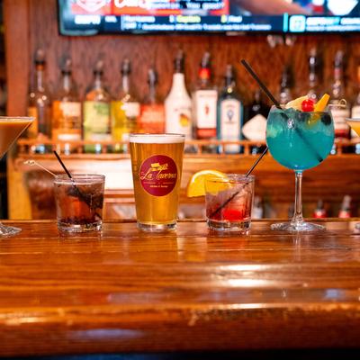 A wooden bar counter displays six colorful cocktails with garnishes and a beer.