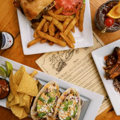 Table spread featuring a burger with fries, tacos with tortilla chips, and a plate of wings.