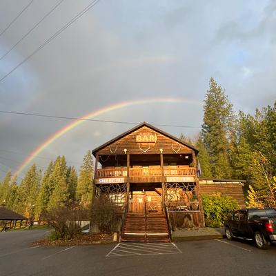 Wooden bar building under a rainbow, surrounded by pine trees.