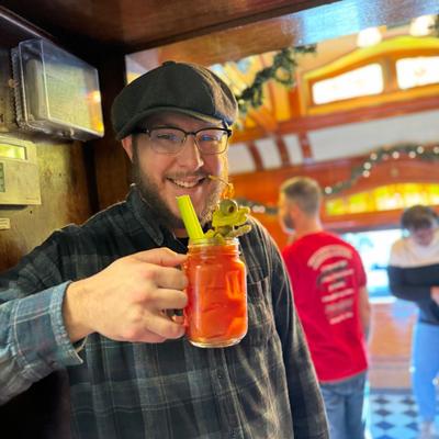 A staffer smiles, holding a red cocktail with a celery stick in a cozy bar.