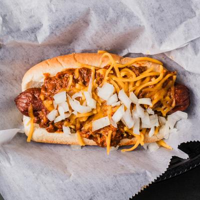 A hot dog with chili, shredded cheddar, and onions, sits on a parchment paper.