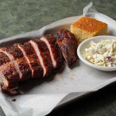 A plate of sliced barbecue ribs served with cornbread and coleslaw.