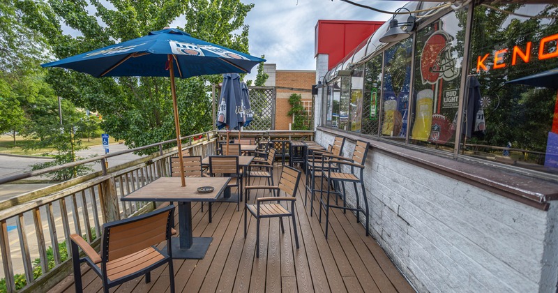 Outdoor patio of a bar with wooden tables and chairs, blue and gray umbrellas, surrounded by trees