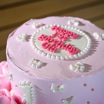 Light pink birthday cake decorated with small ribbon bows, pearls, and a central plaque.