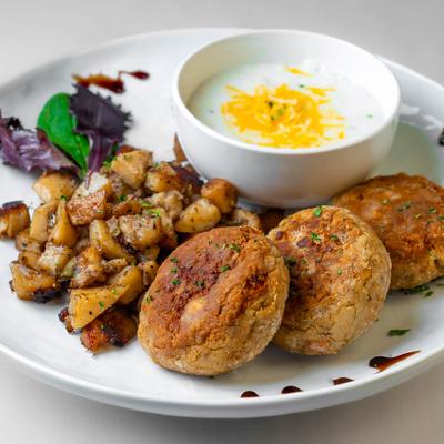Crab cakes served with seasoned potatoes and a bowl of creamy soup topped with cheese.