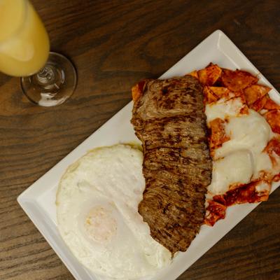 Carne asada steak served alongside fried eggs, chilaquiles, and a mimosa.