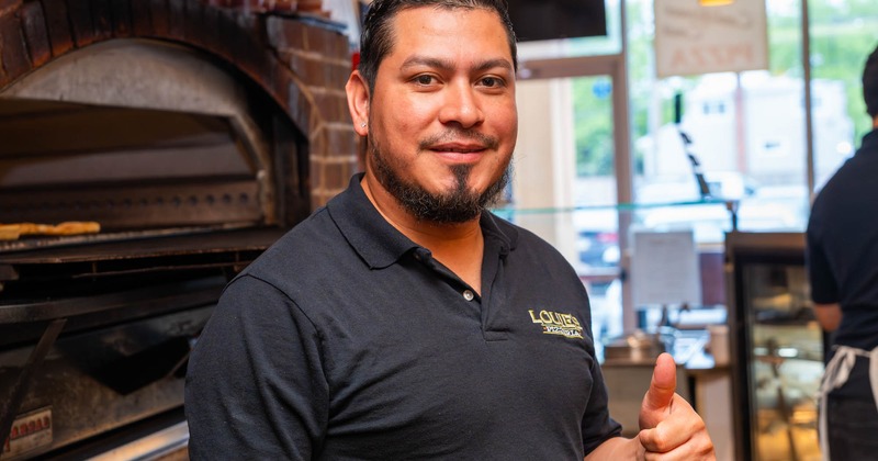 An employee smiling and giving a thumbs-up, standing near a brick oven